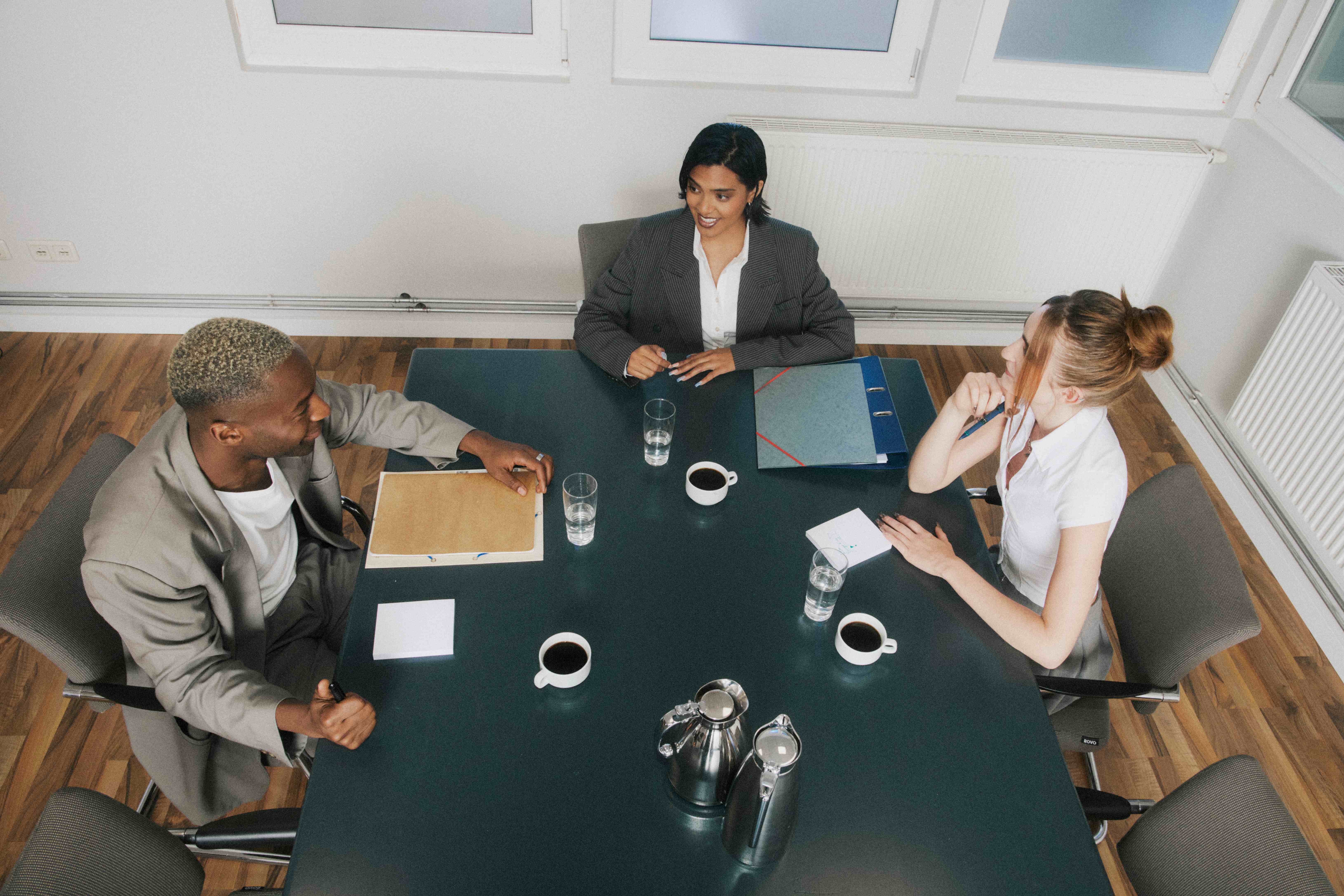 Overhead of three colleagues at a dark boardroom table with coffee, water, and folders — The Ad Spend brand image
