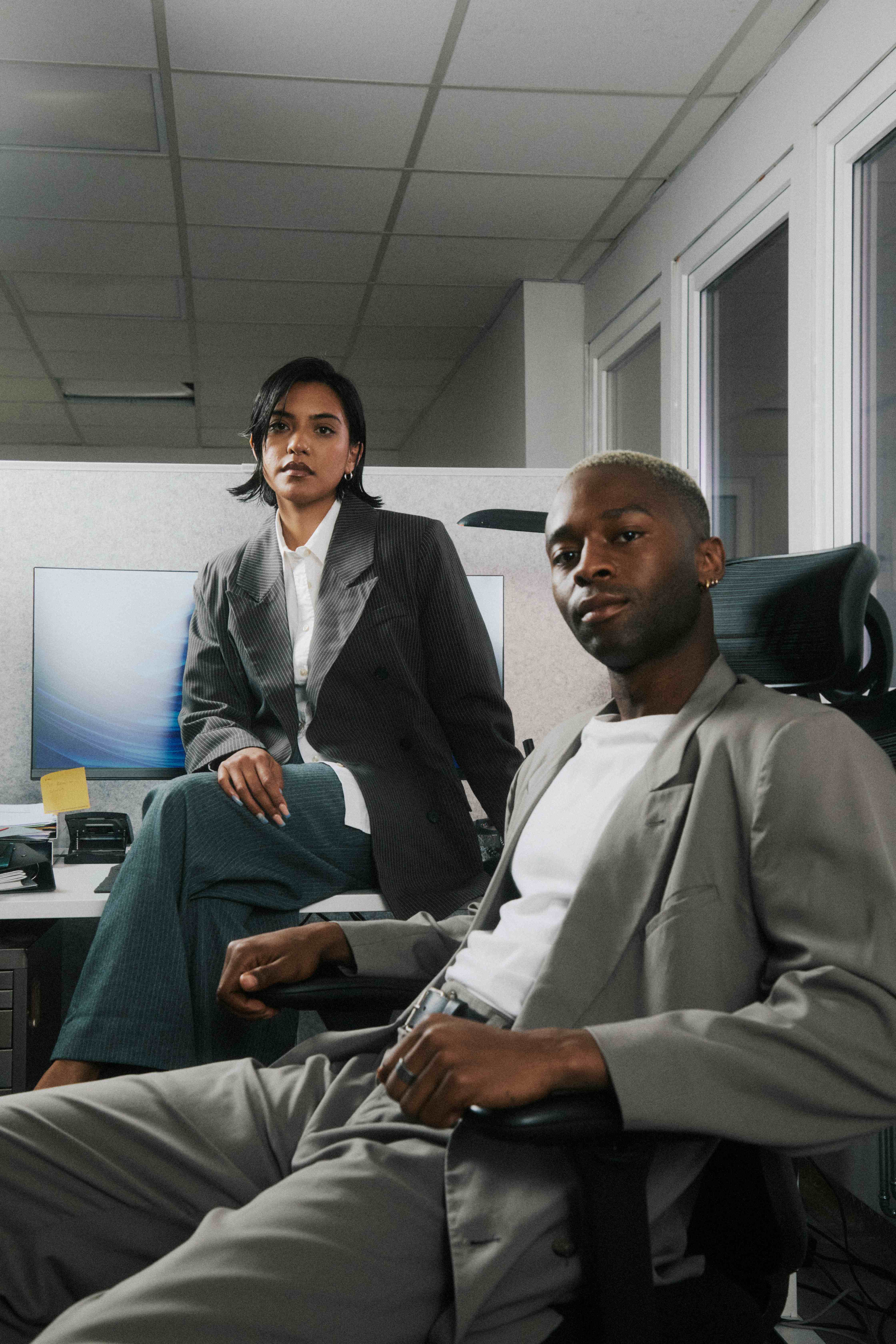 Two colleagues posing confidently to camera — woman on a desk edge, man in a chair — The Ad Spend brand image