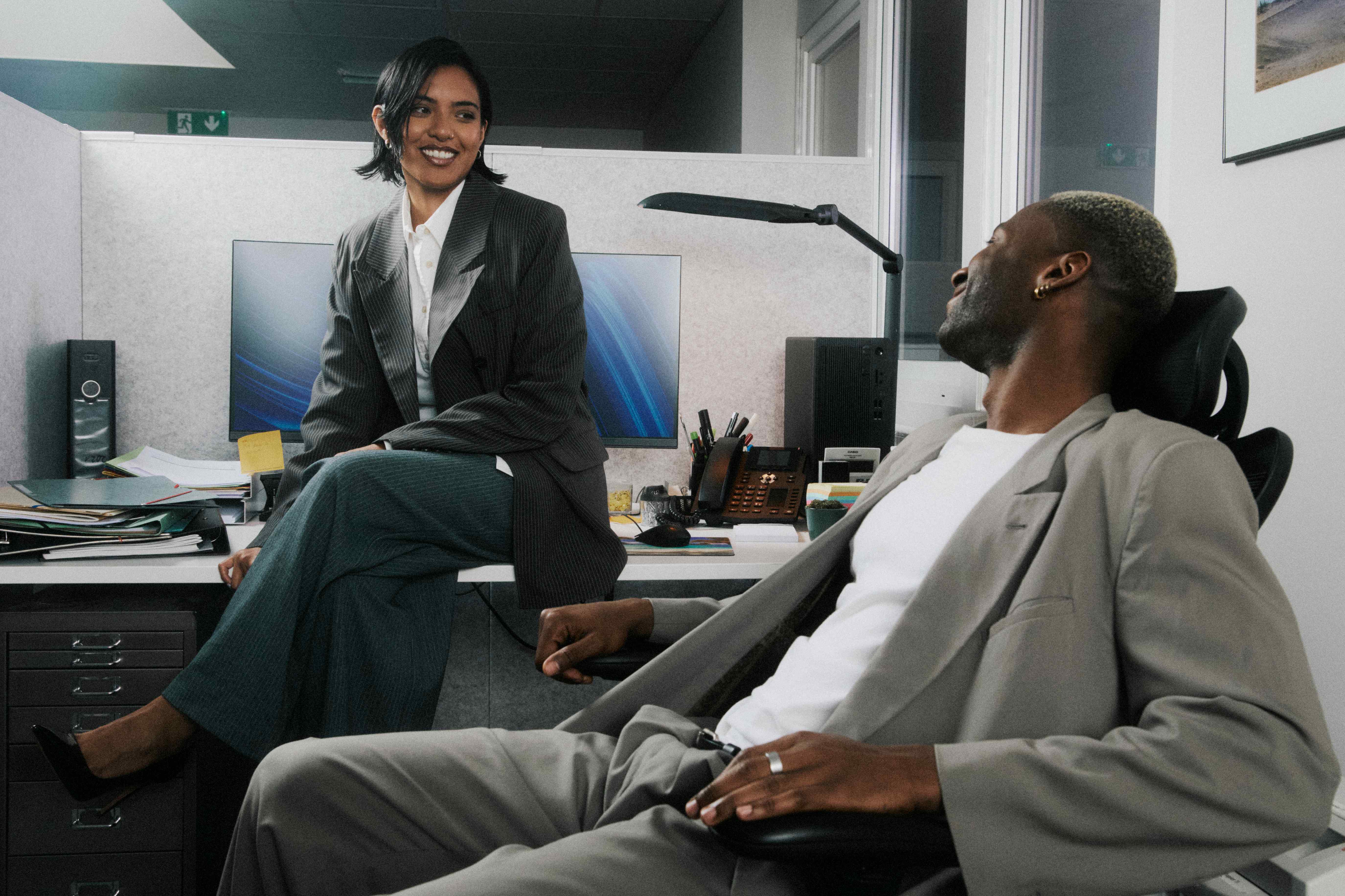 Woman sitting on a desk edge laughing with a man reclining in a chair — The Ad Spend brand image