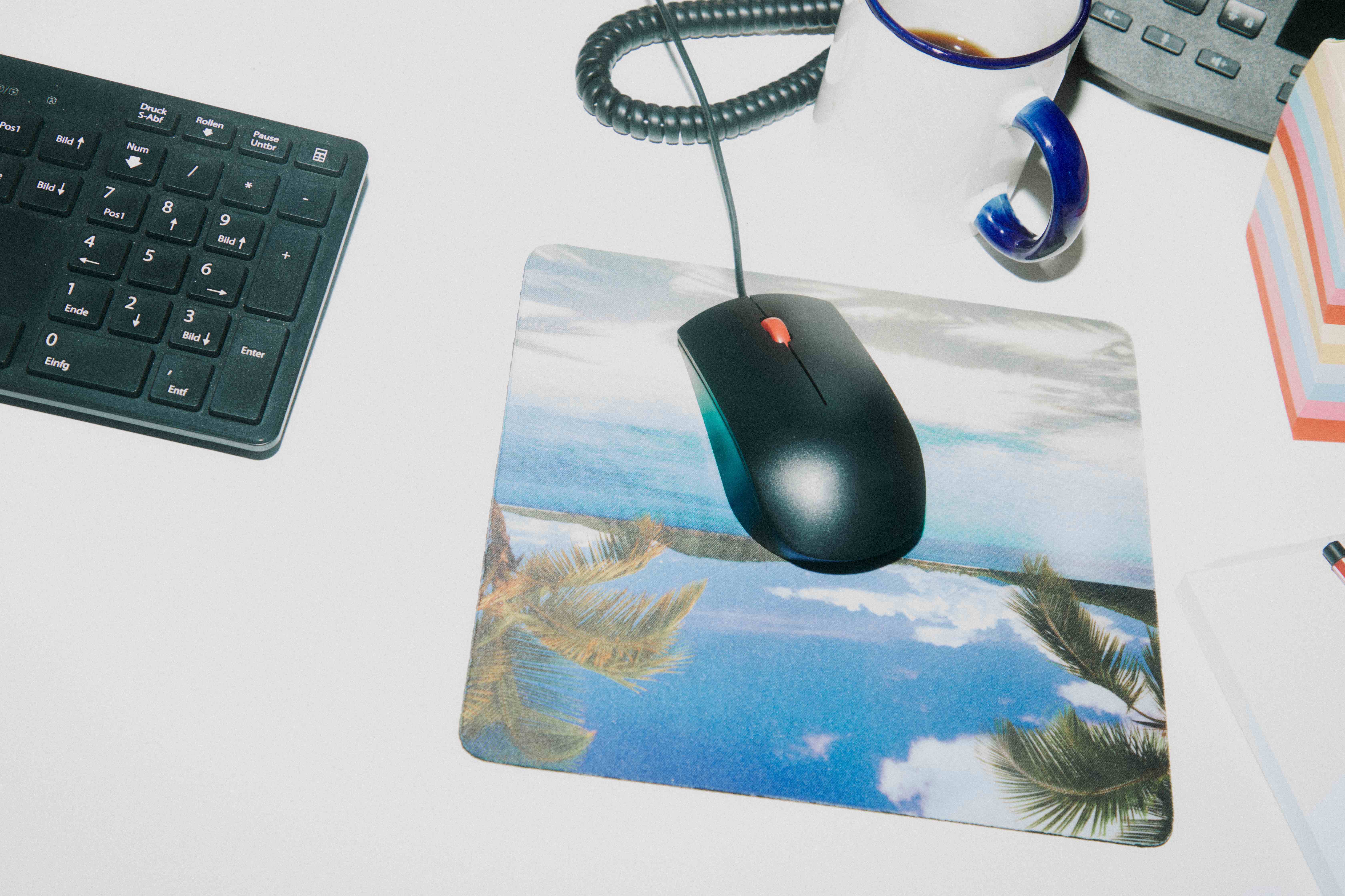Top-down desk with keyboard, mouse on a tropical-palm mousepad, and a blue-rimmed coffee mug — The Ad Spend brand image