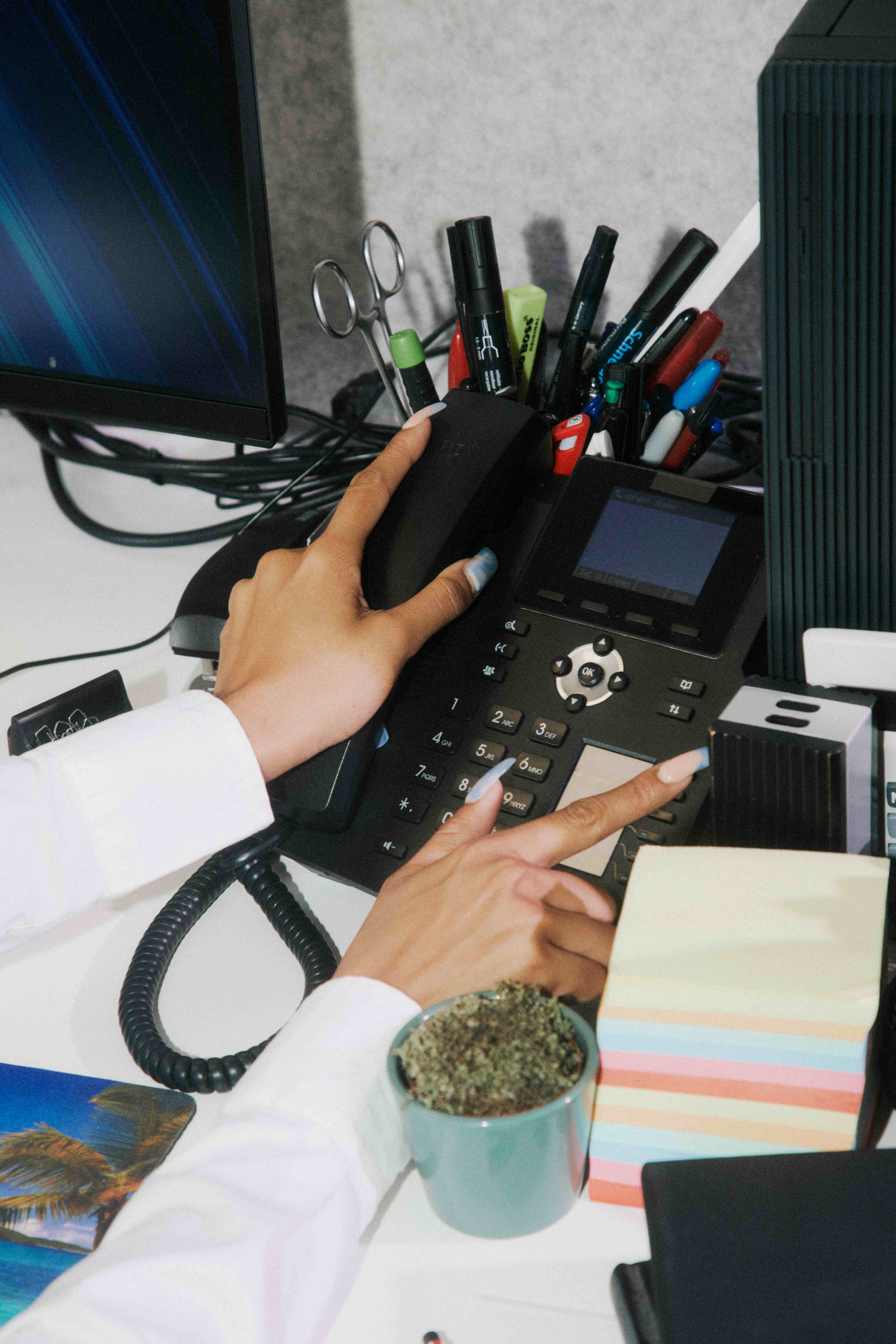 Overhead of hands using a black desk phone; a pen pot with scissors and markers on a palm-tree mousepad — The Ad Spend brand image