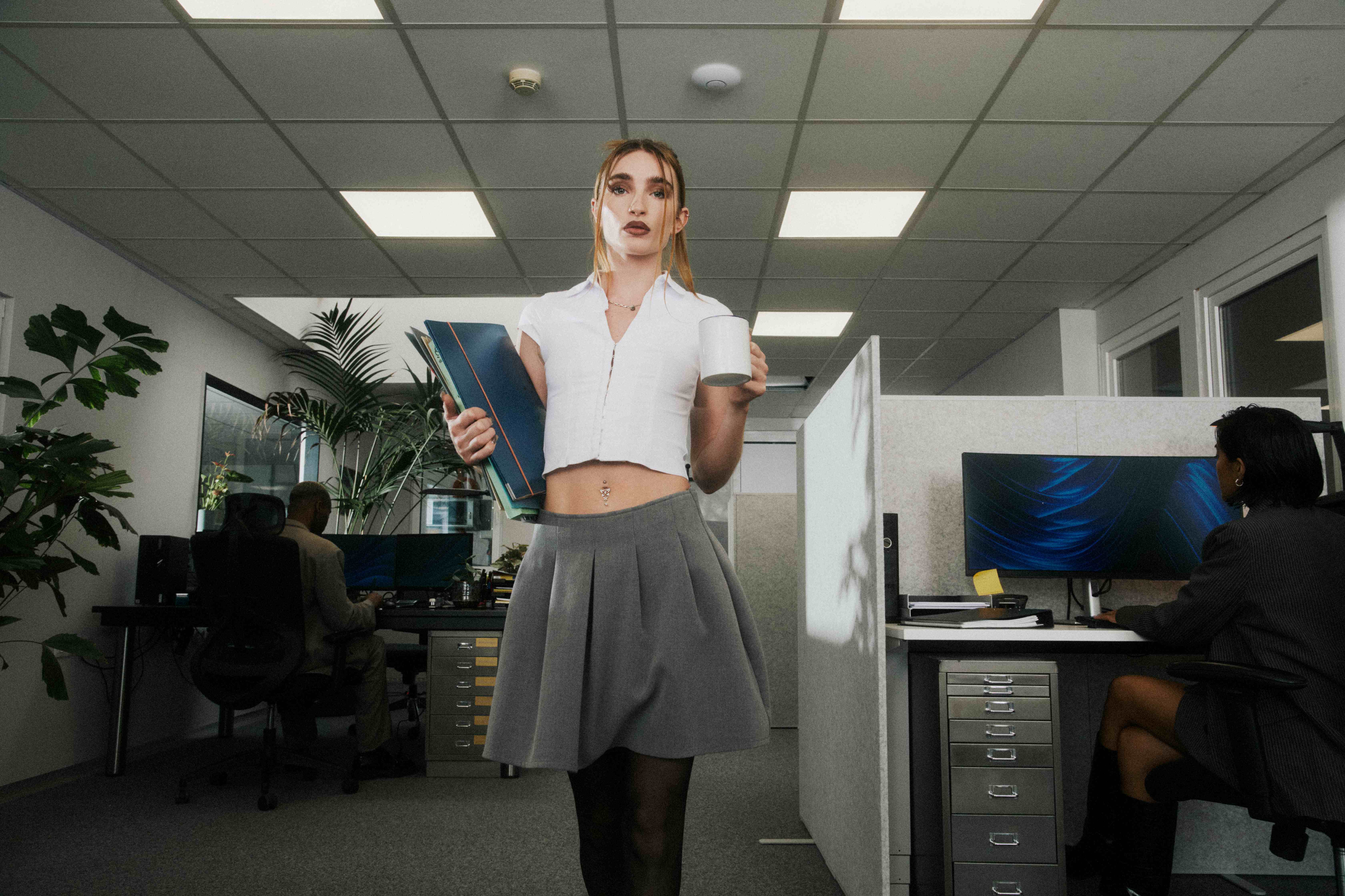 Woman in a white top and grey pleated skirt holding a binder and mug, facing camera; colleagues working behind — The Ad Spend brand image