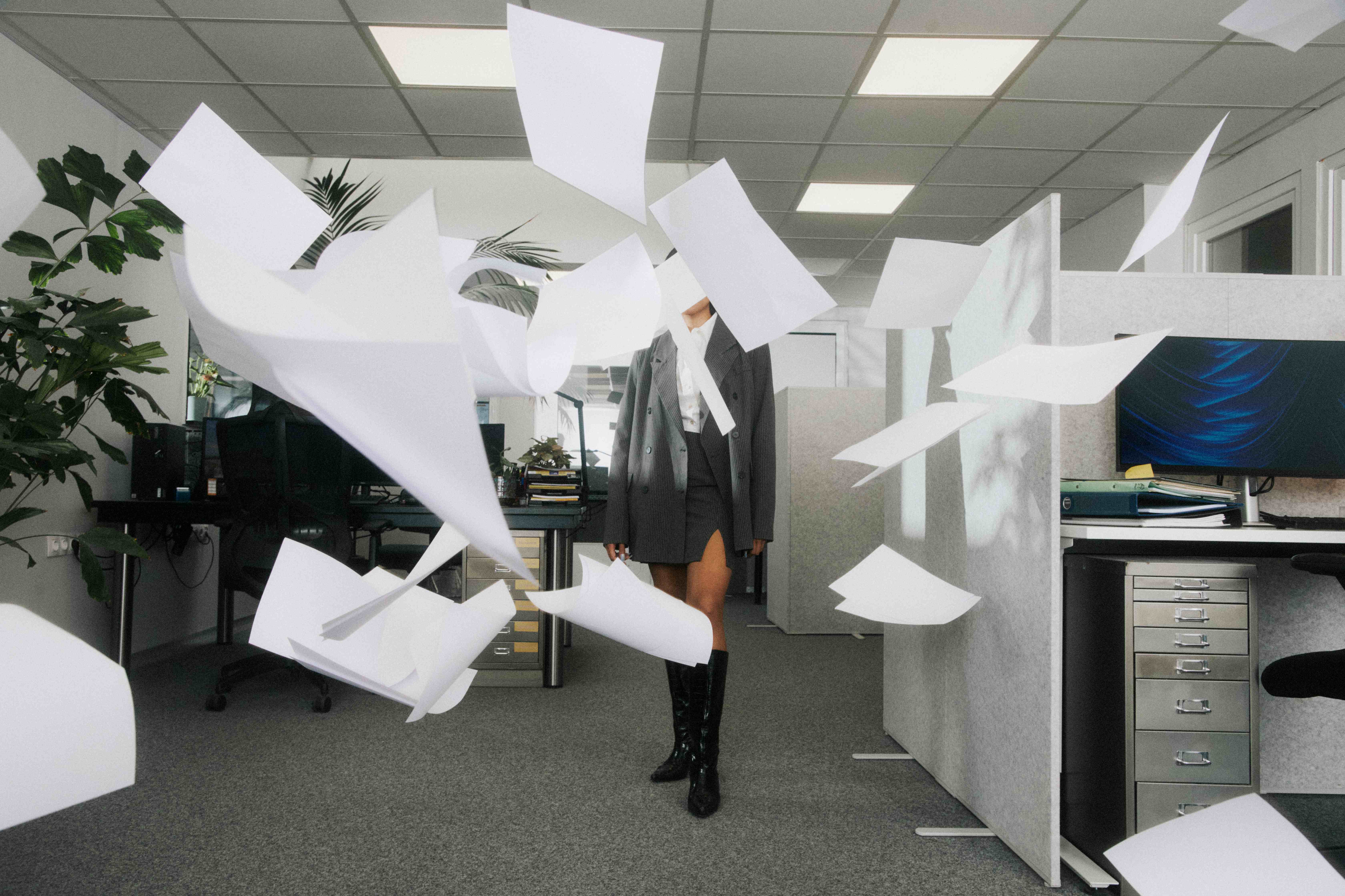 Composed figure in oversized grey pinstripe amid a flurry of flying paper in a cubicle office, face obscured by paper — The Ad Spend brand image
