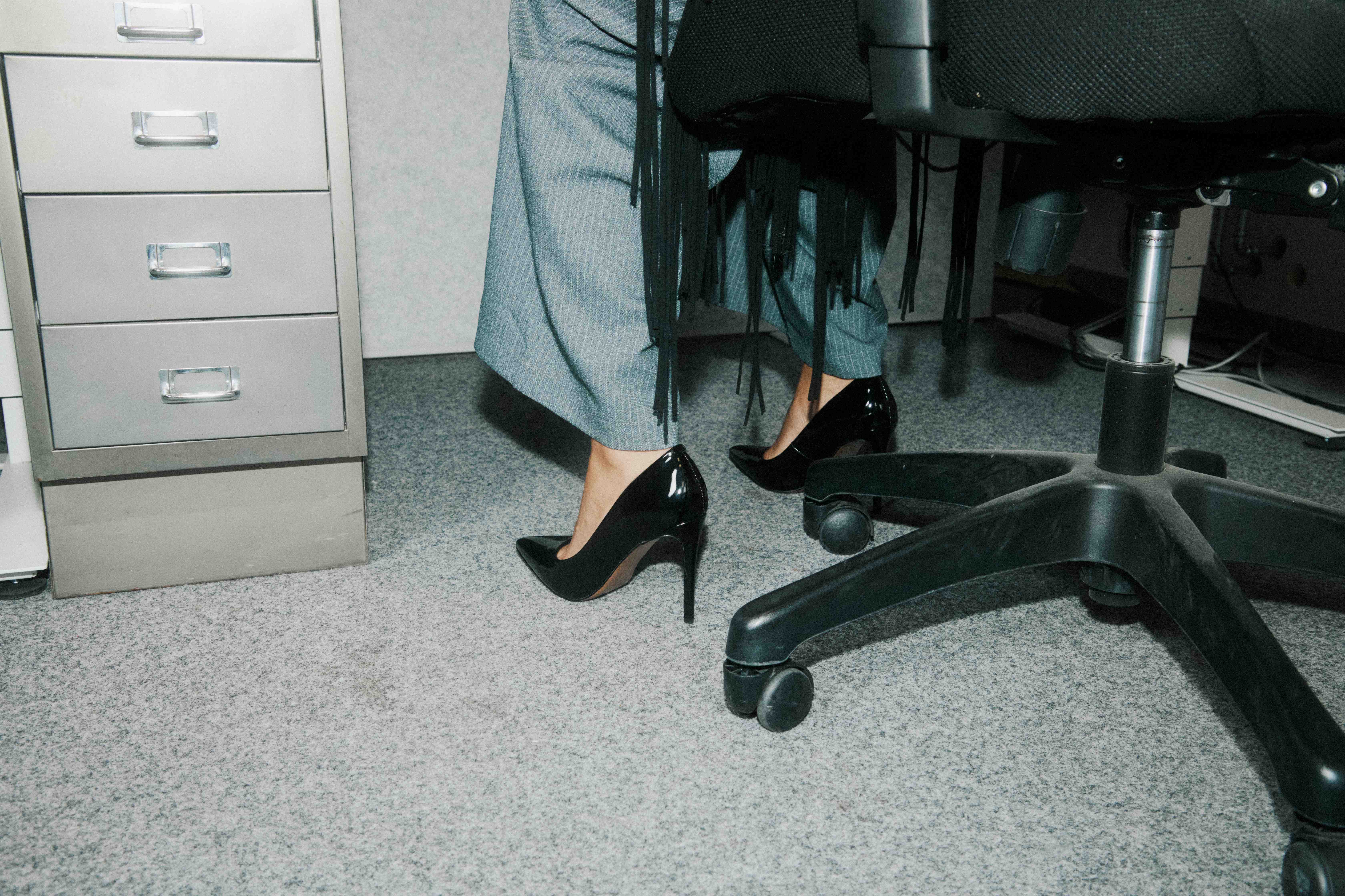 Low-angle of black patent heels beside an office chair's wheel base and a metal filing cabinet — The Ad Spend brand image