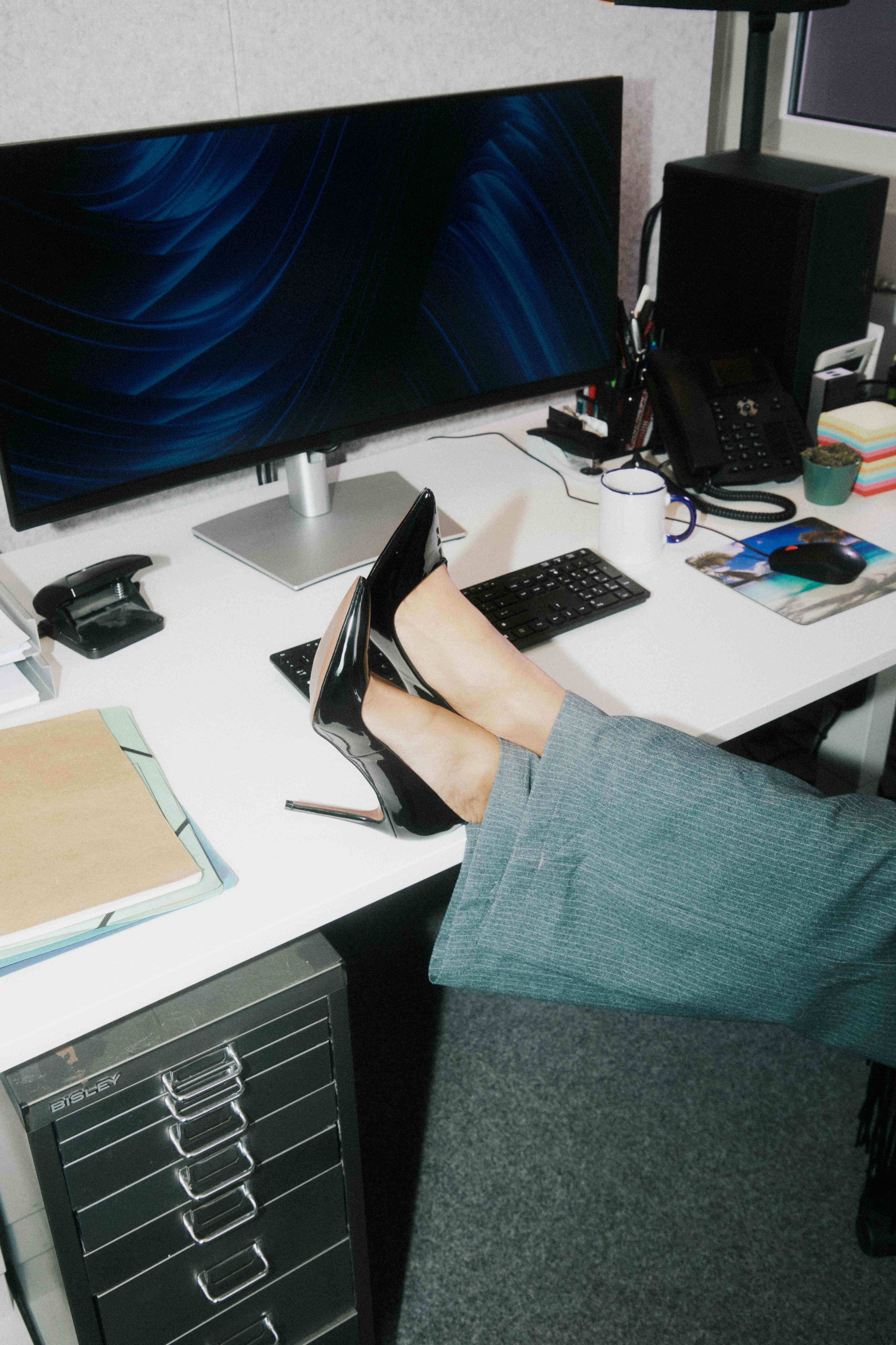 Black patent heels propped on the desk beside the keyboard; kraft folder and a blue-wallpaper monitor — The Ad Spend brand image