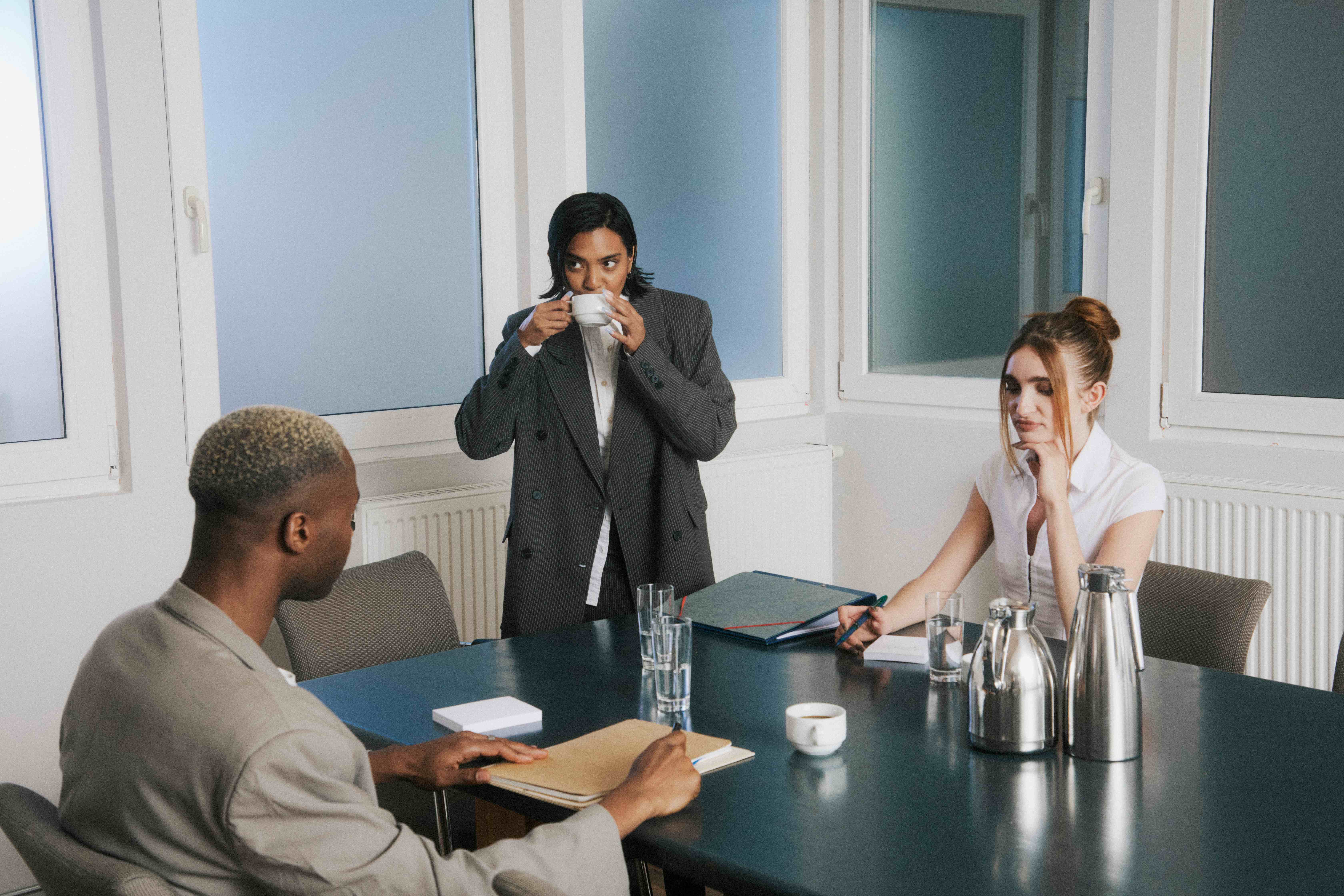 Meeting room: a woman standing sipping coffee while two colleagues sit at the table — The Ad Spend brand image